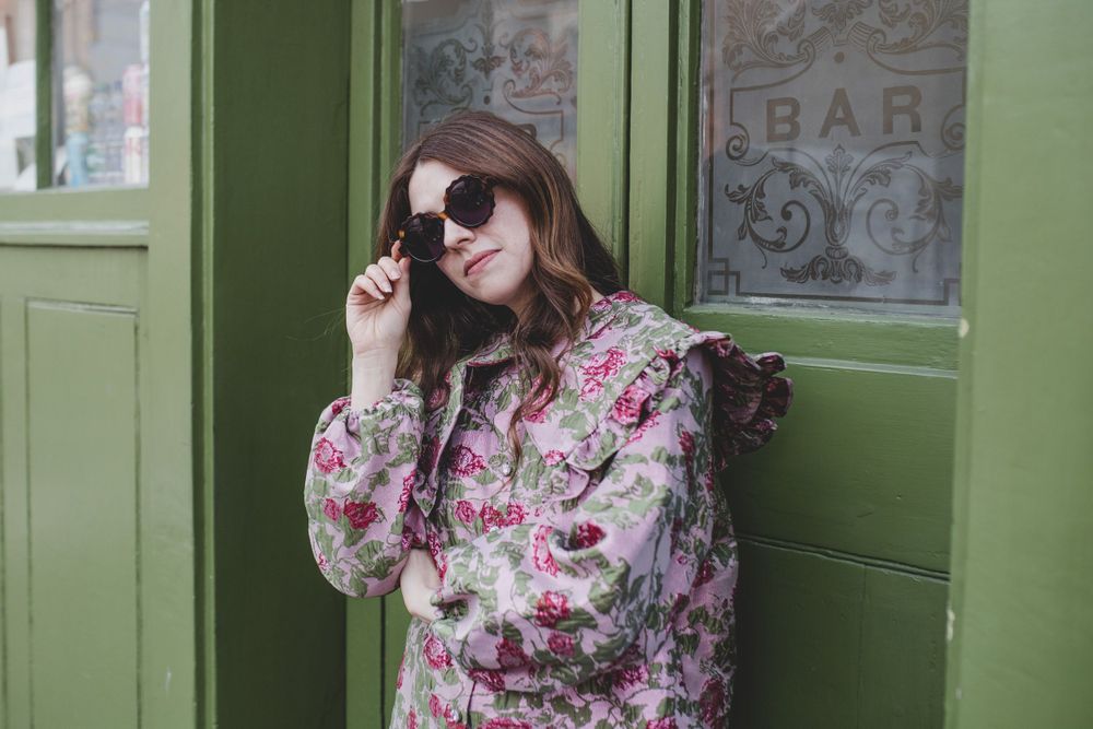 Vintage-inspired fashion pose with sunglasses outside green shopfront.