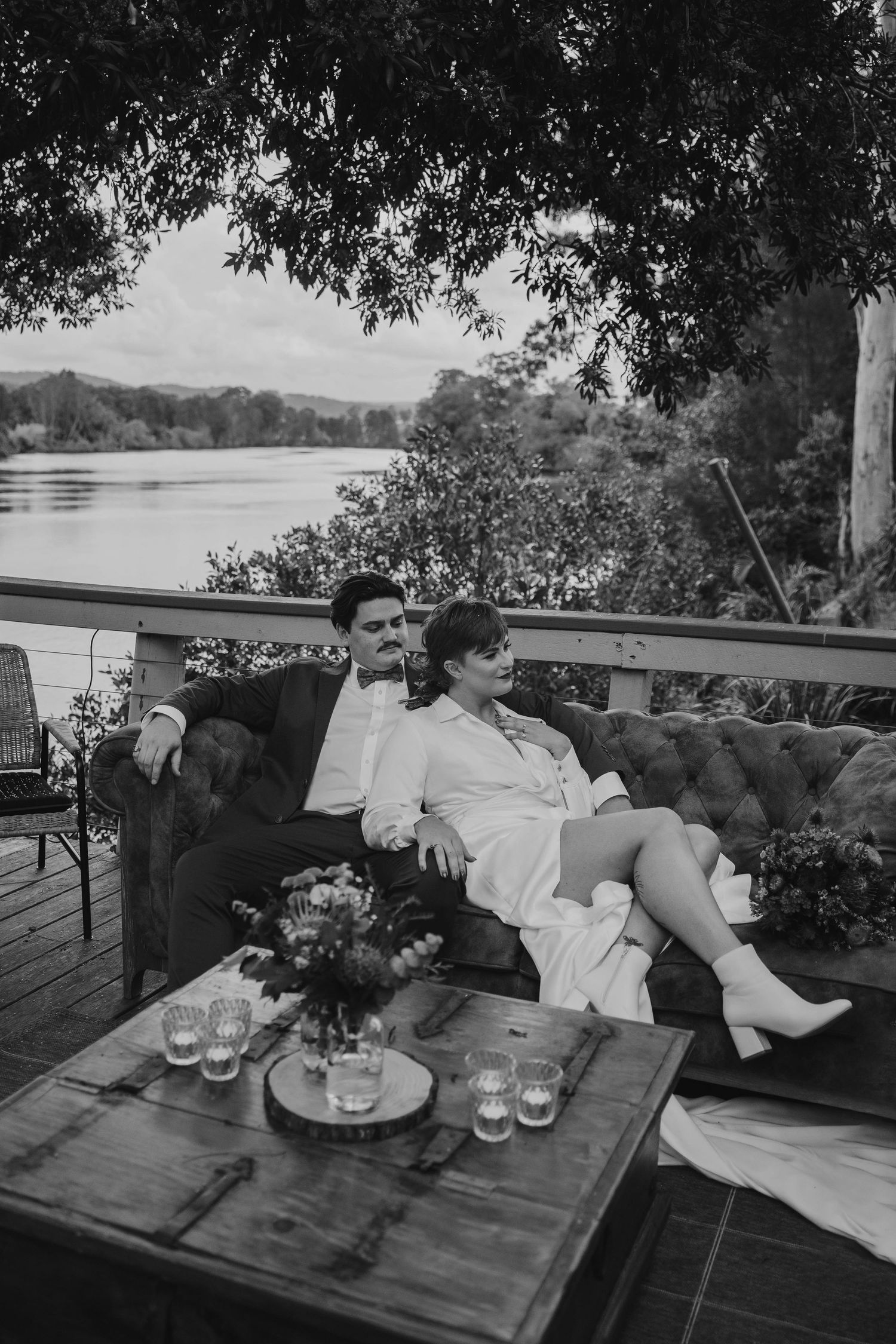 Black and white wedding photo of couple relaxing on outdoor deck by lake.