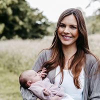 Eine Mutter hält ihr Baby auf dem Arm und lächelt in die Kamera. Sie stehen vor einer grünen Natur-Kulisse.