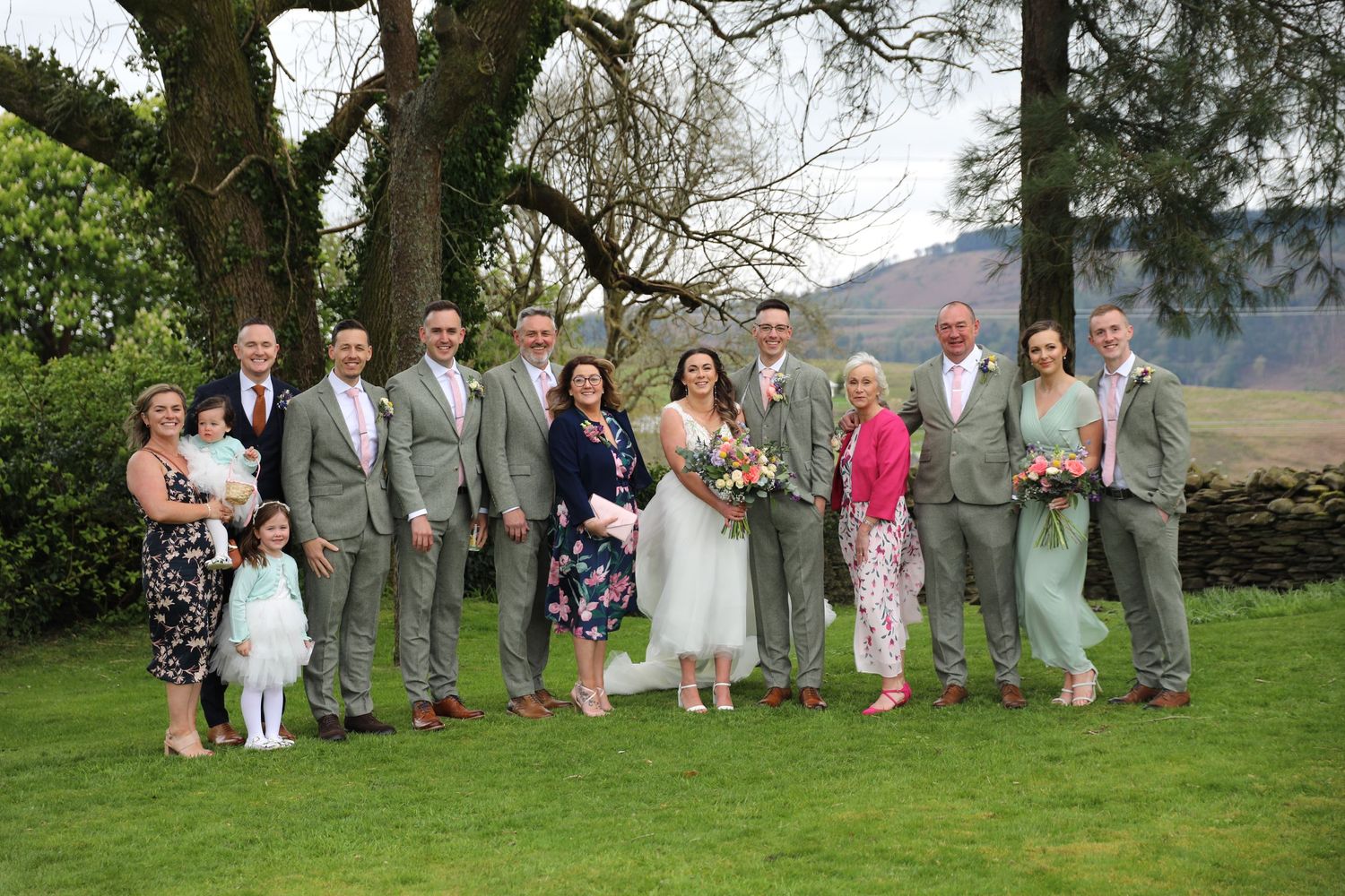 Wedding party in grey and white attire poses on green lawn with mountain backdrop.