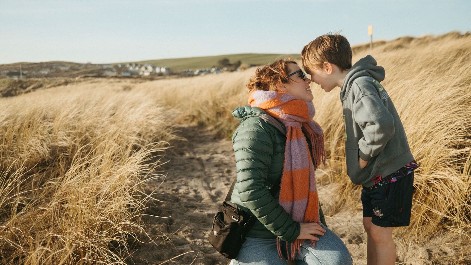 Two figures embracing on a coastal path with tall golden grass and ocean views in the background.