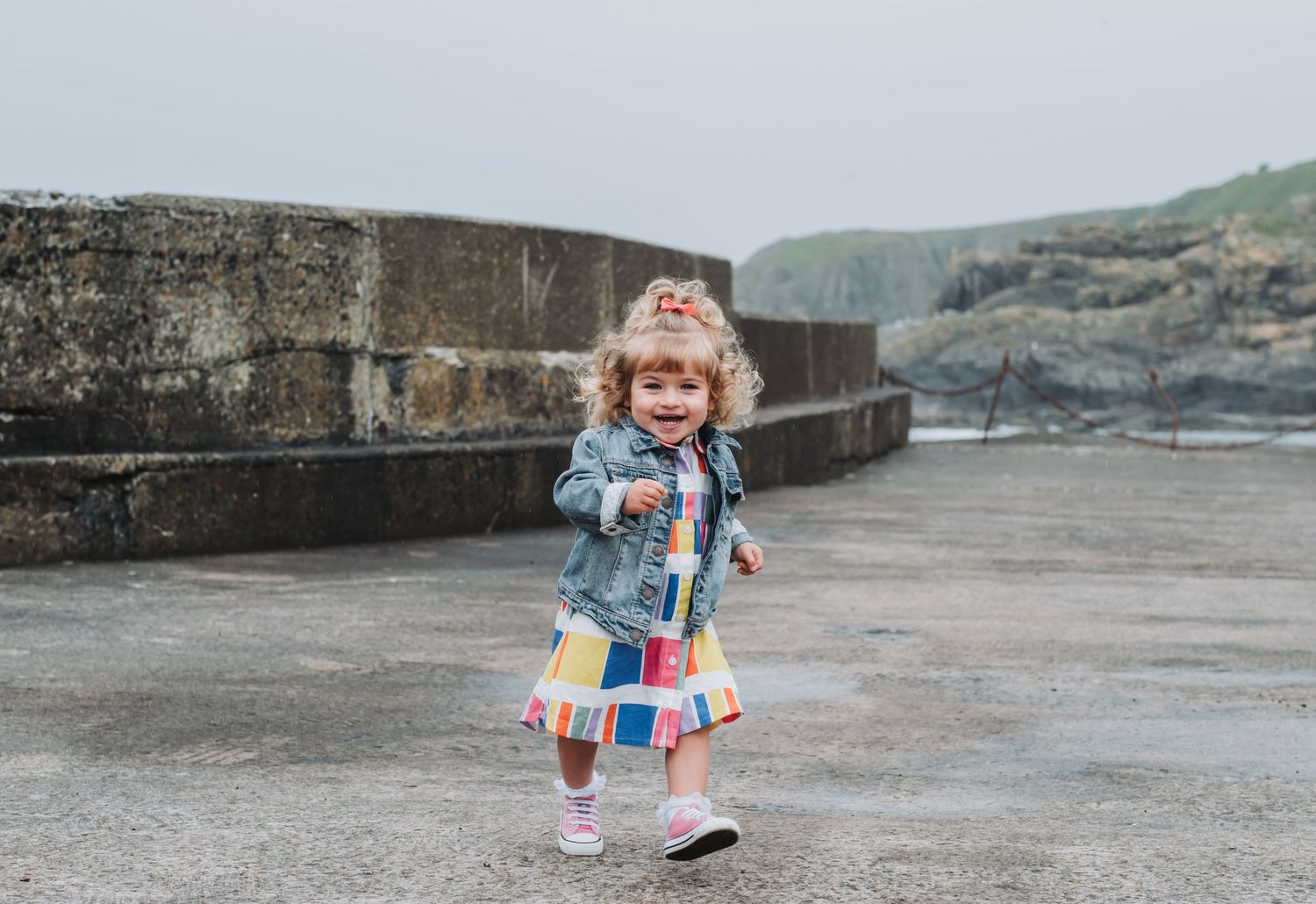 A cheerful toddler girl in a colorful dress and denim jacket running along a coastal harbor path.