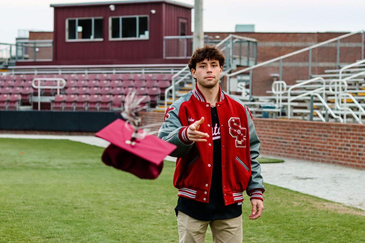 Student wearing red varsity jacket stands on sports field with stadium seats in background.
