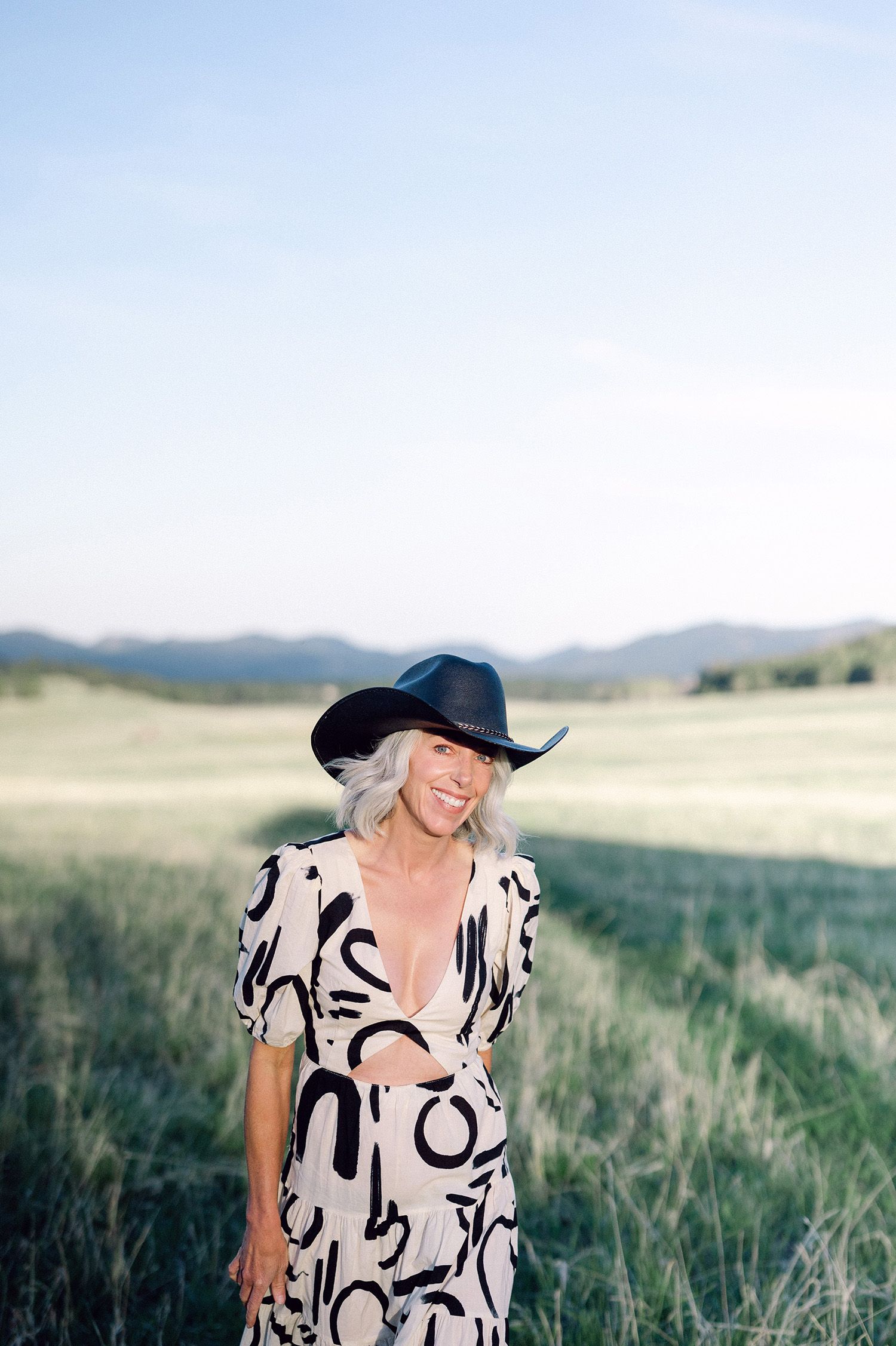 Portrait of woman in black sweater and cream fedora hat with mountain backdrop.