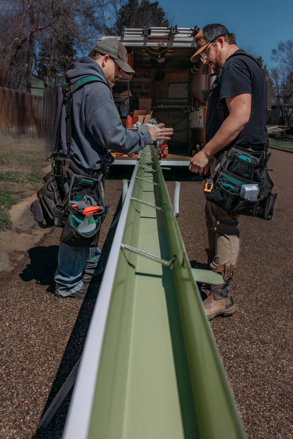 Three people inspect and work on a large green surfboard or board resting on sawhorses outdoors under a blue sky.