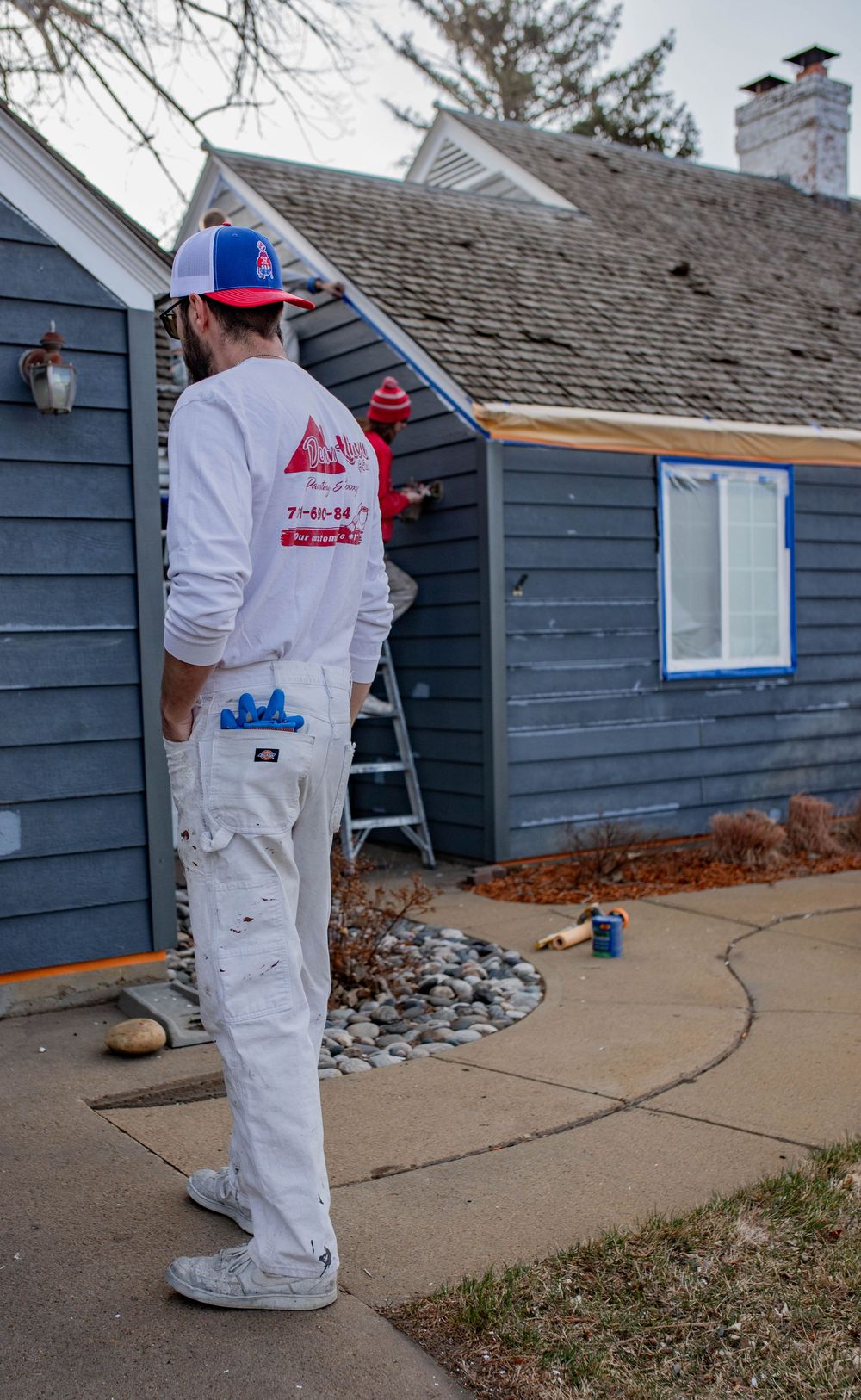 Painters in white coveralls working on exterior blue house siding by adding protective tape along concrete path.