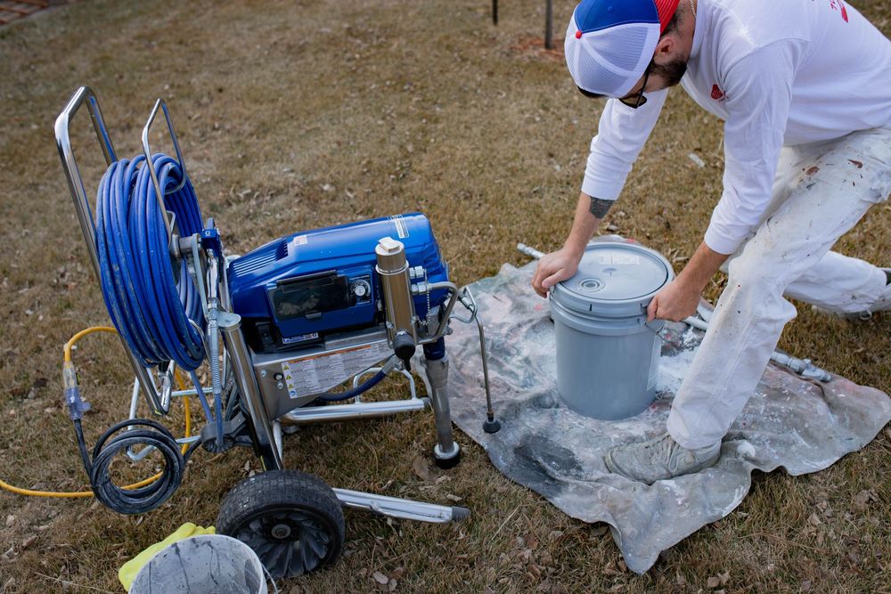 Professional in protective suit applying residential foundation waterproofing with blue equipment cart and tools nearby.