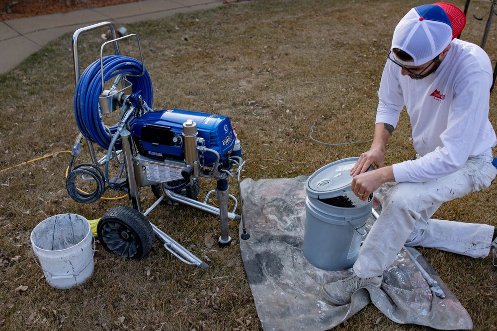 A painter prepares equipment and materials with a professional airless paint sprayer and supplies on the ground.