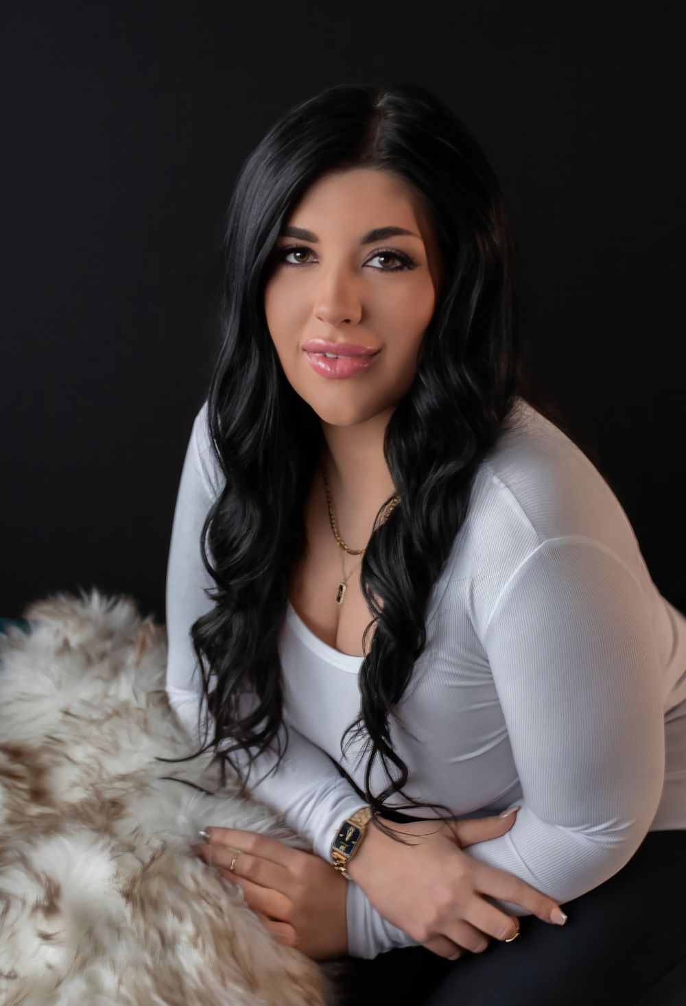A woman in a white fitted top poses with a fur throw against a dark background.