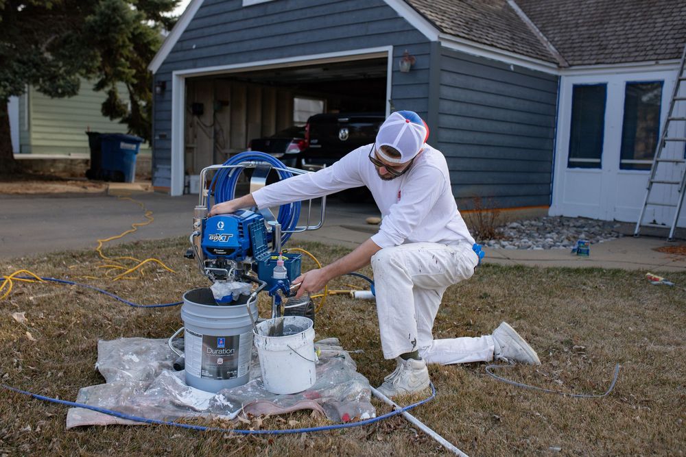 A house painter sets up a paint sprayer machine on protective tarps in front of a grey residential garage.