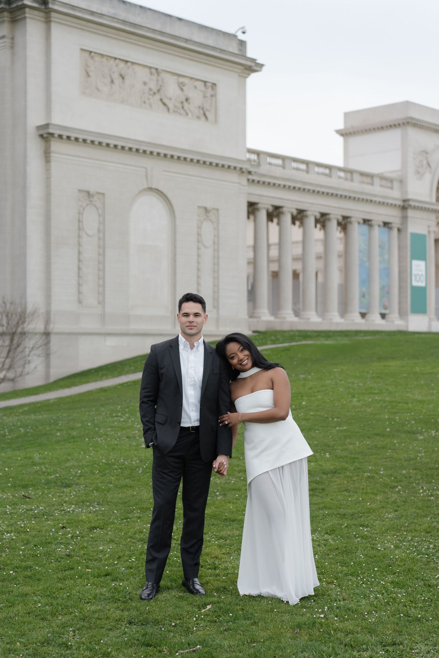 A couple in wedding attire poses on a grassy lawn in front of a grand neoclassical building with white columns.