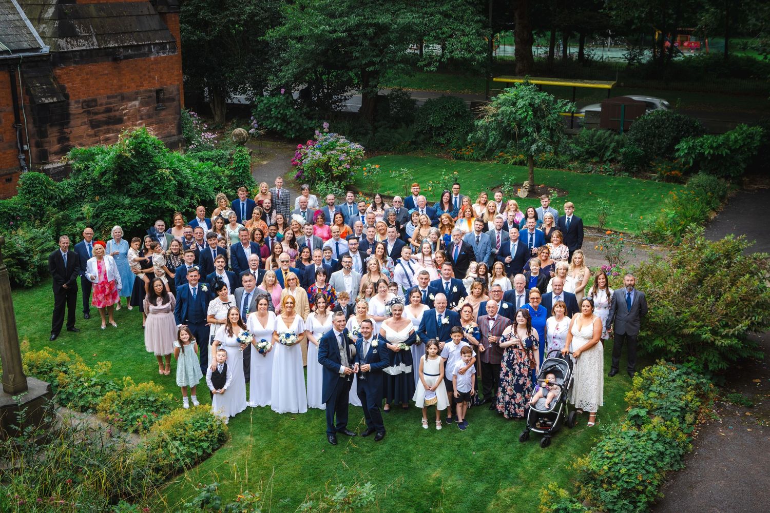 Large wedding party poses together in a lush garden setting with brick building backdrop.