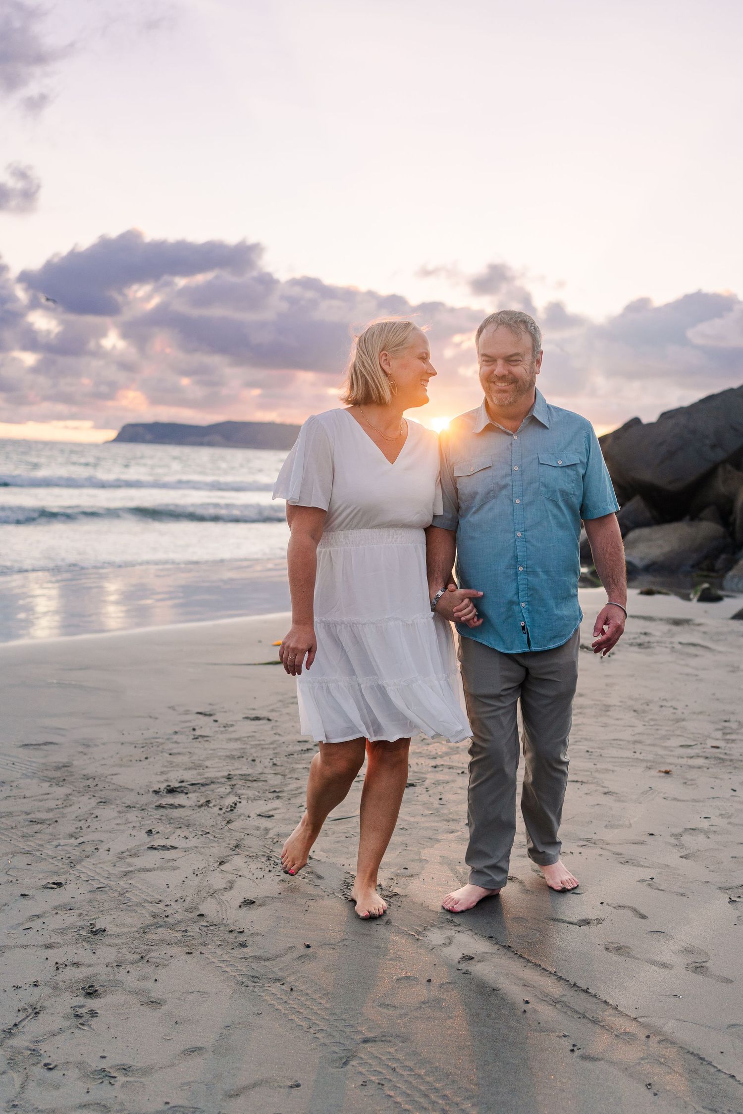 Couple strolling on beach at sunset with sun peeking between them.