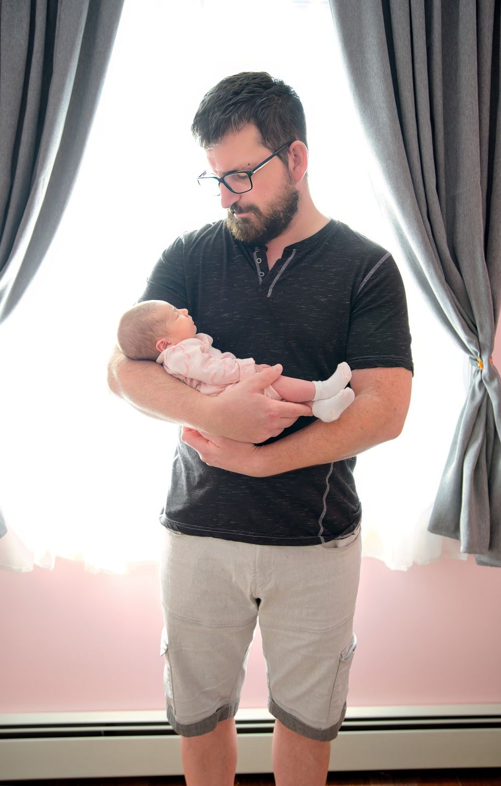 A bearded man in glasses and casual attire tenderly holds a sleeping newborn baby in front of gray curtains.