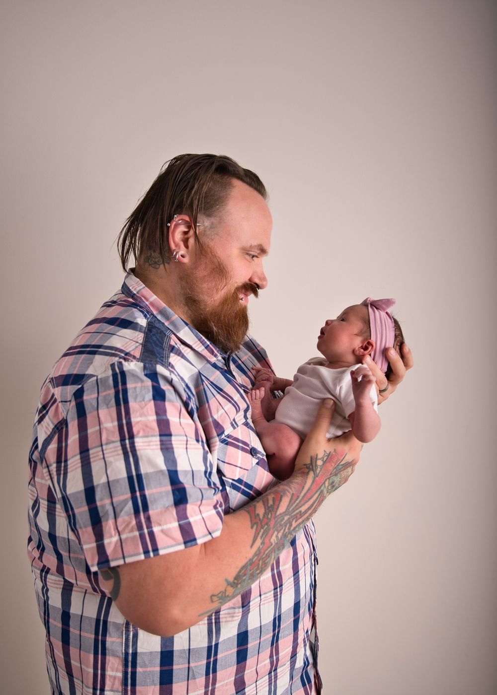 Bearded man in plaid shirt holding and gazing lovingly at newborn baby against neutral background.