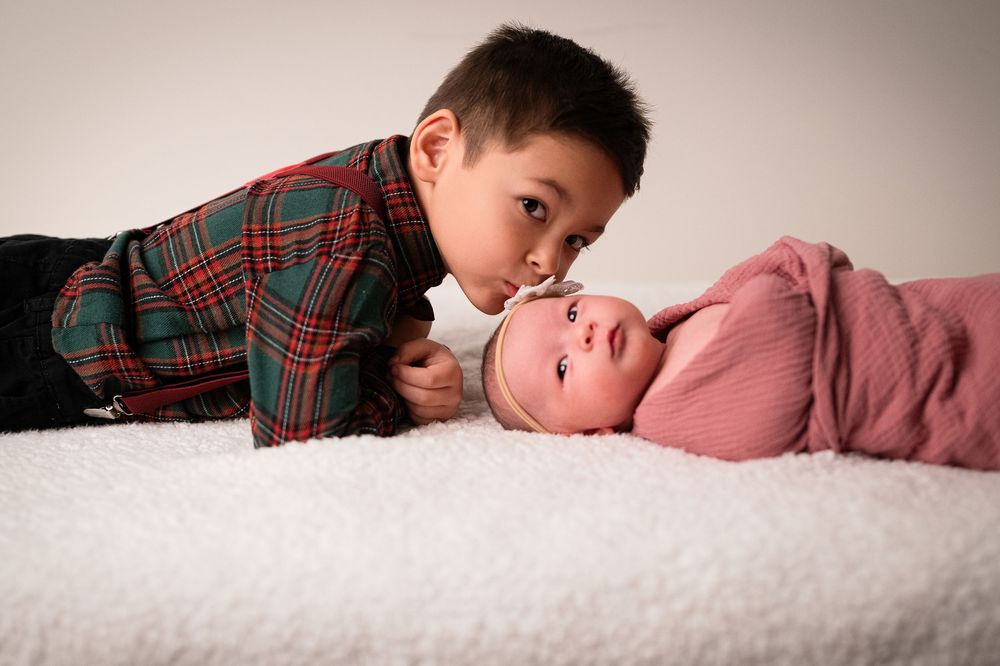 A young child in a plaid shirt gently interacts with a sleeping baby on white bedding.