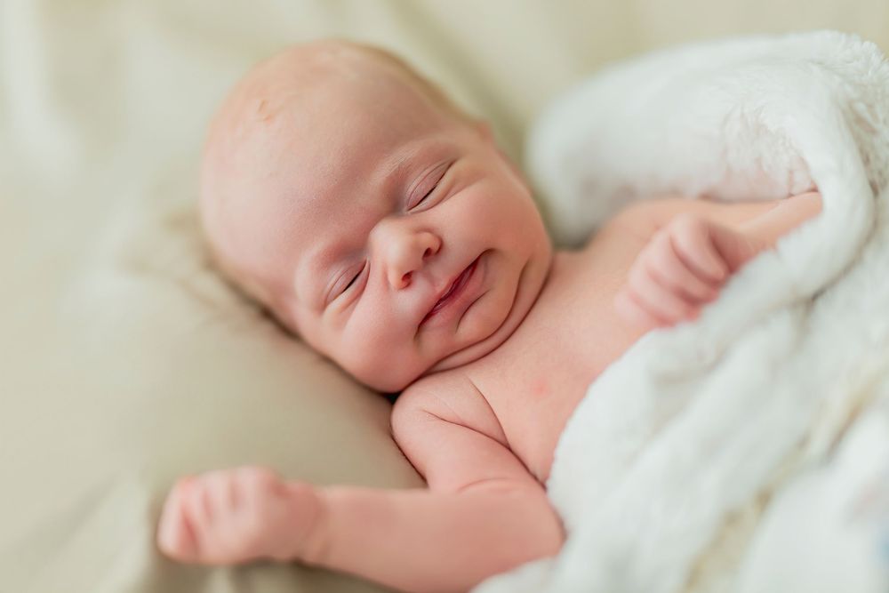 Peaceful sleeping newborn baby with rosy cheeks wrapped in soft white blanket against cream background.