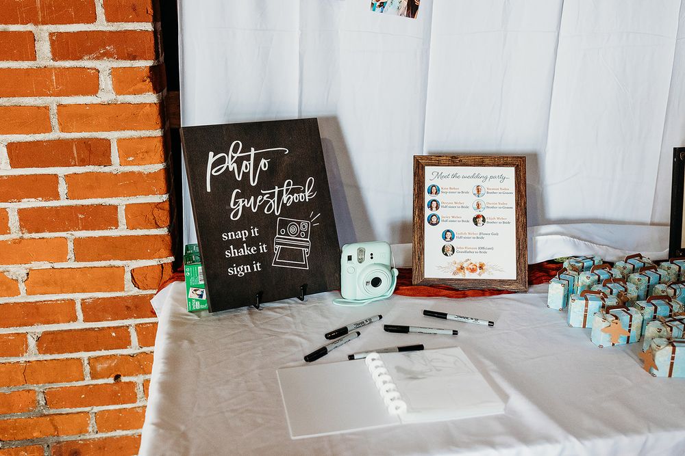 Handwritten wedding signage and guest book setup on white table against brick wall with decorative mint accents.
