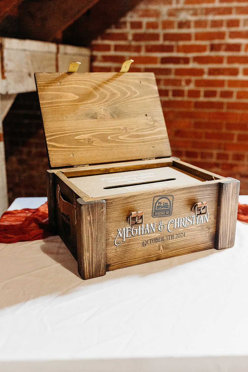 Rustic wooden wedding card box with engraved names sits open on white tablecloth against brick wall backdrop.