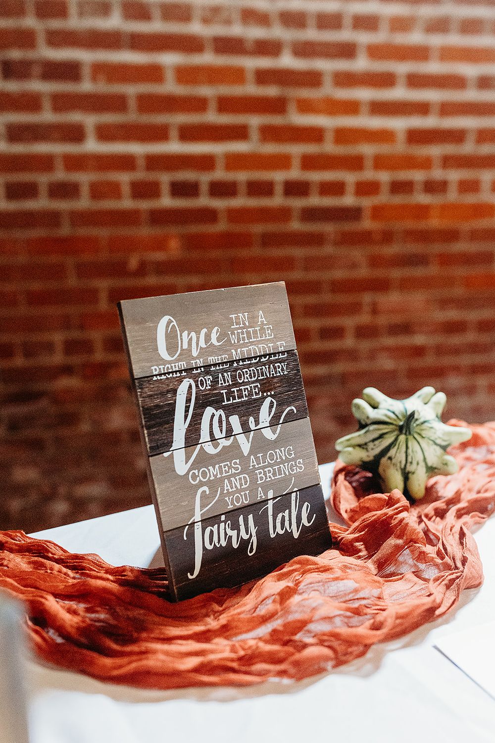 A rustic wedding sign with romantic text sits on a coral-colored fabric next to a small succulent against a brick wall.