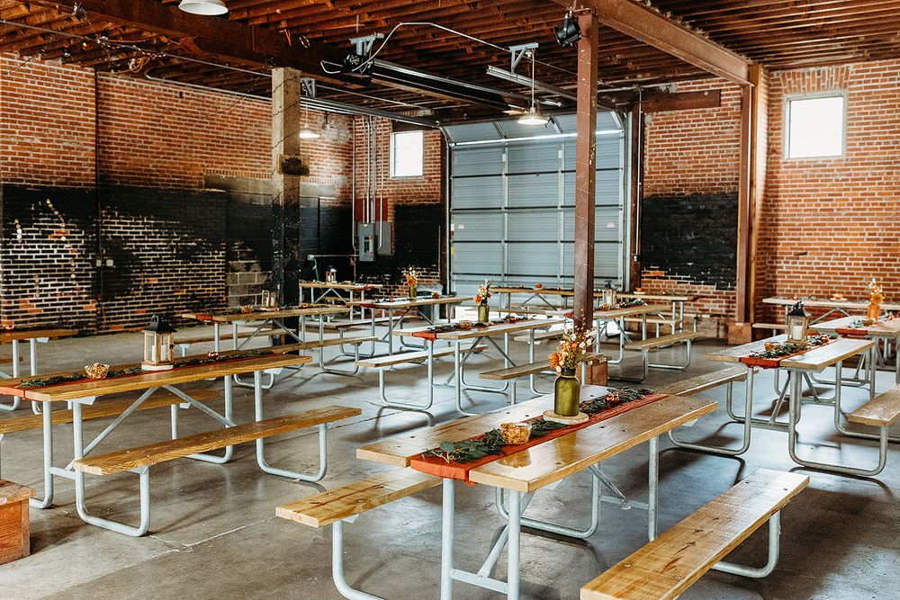 Industrial chic dining hall with white picnic tables, exposed brick walls, and wooden ceiling beams in a rustic event space.