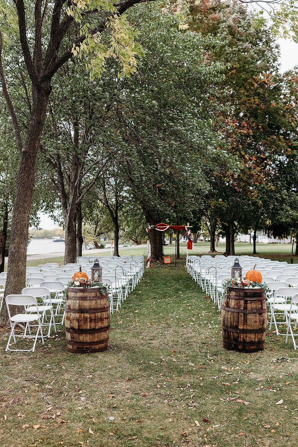 Rustic outdoor wedding ceremony setup with white chairs lined up between wooden barrels decorated with autumn pumpkins.