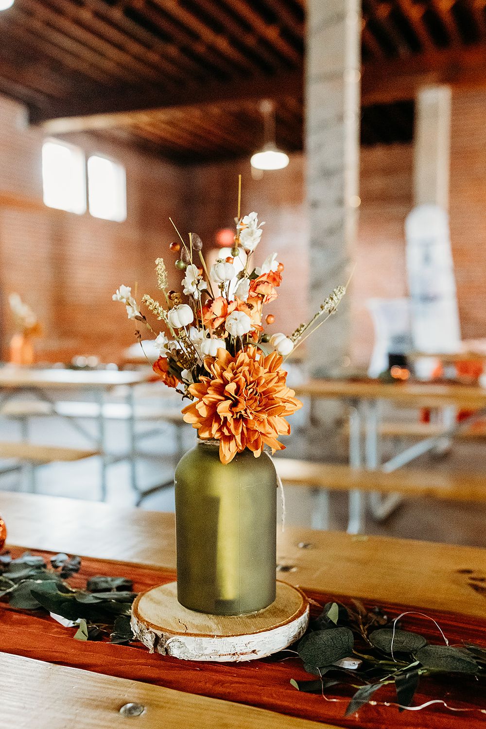 A rustic autumn centerpiece featuring orange flowers and white blooms in a gold-painted vase on a wooden table in a barn setting.