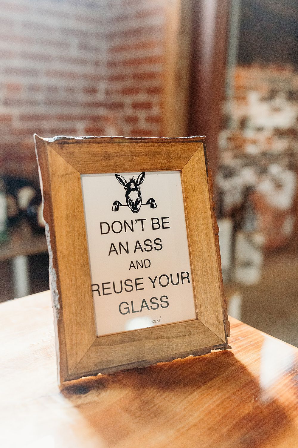 A framed sign with wooden border reads 'DONT BE AN ASS AND REUSE YOUR GLASS' with a decorative donkey logo on top.