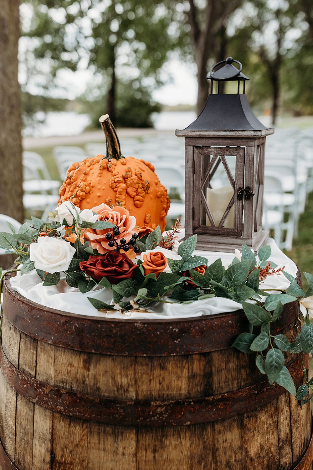Fall wedding decor featuring a rustic wooden barrel topped with an orange pumpkin, lantern, and floral arrangements.
