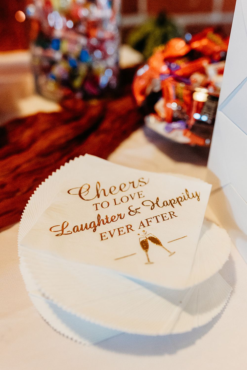 White wedding napkin with text 'Cheers to Laughter & Happily Ever After' in gold calligraphy on a table with blurred decor.