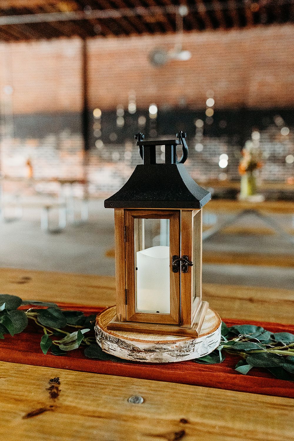 Rustic wooden lantern with glowing candle sits on birch slice base with greenery holiday decor on wooden table.