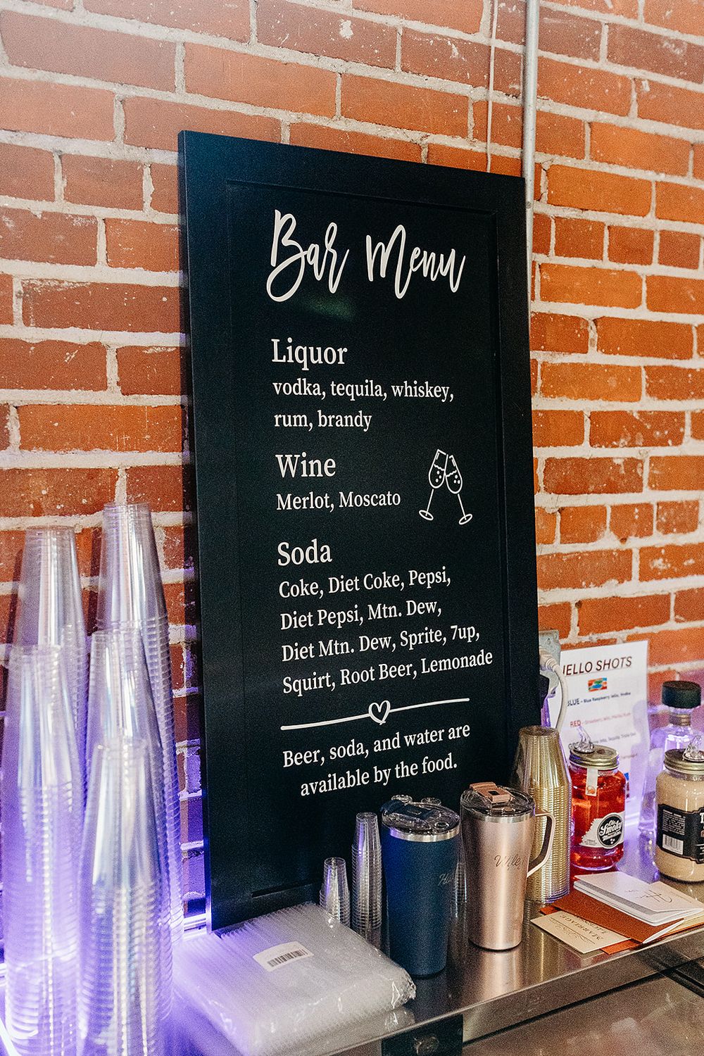 Black chalkboard bar menu displays beverage options including liquor, wine and soda against a brick wall with purple backlit glasses.