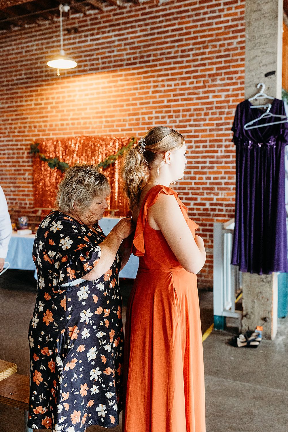 Two people stand near a brick wall in a wedding venue while one helps adjust a bright orange dress as purple gowns hang nearby.
