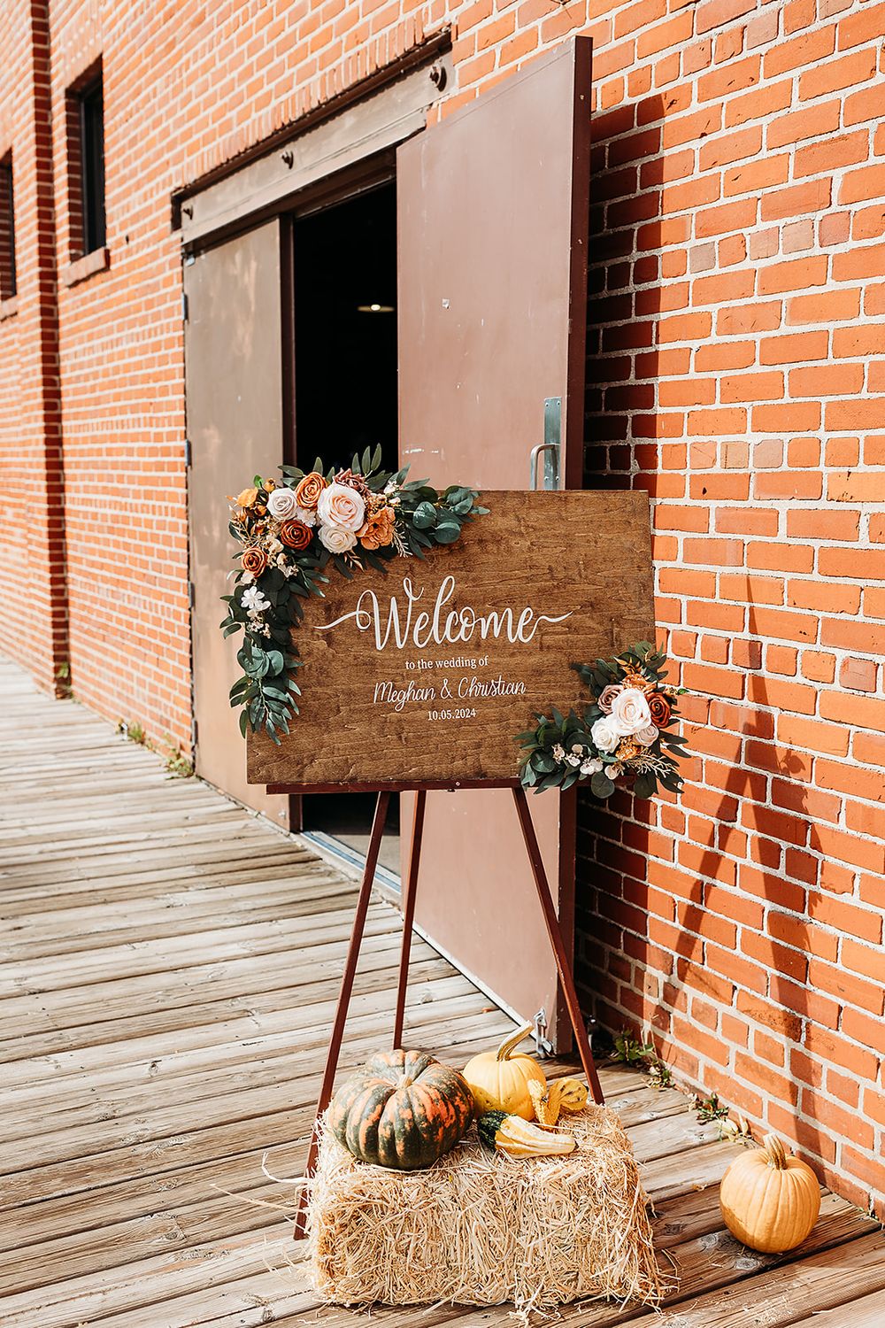 Rustic wooden welcome sign with floral decorations and pumpkins on hay bales against a brick wall at a fall wedding.