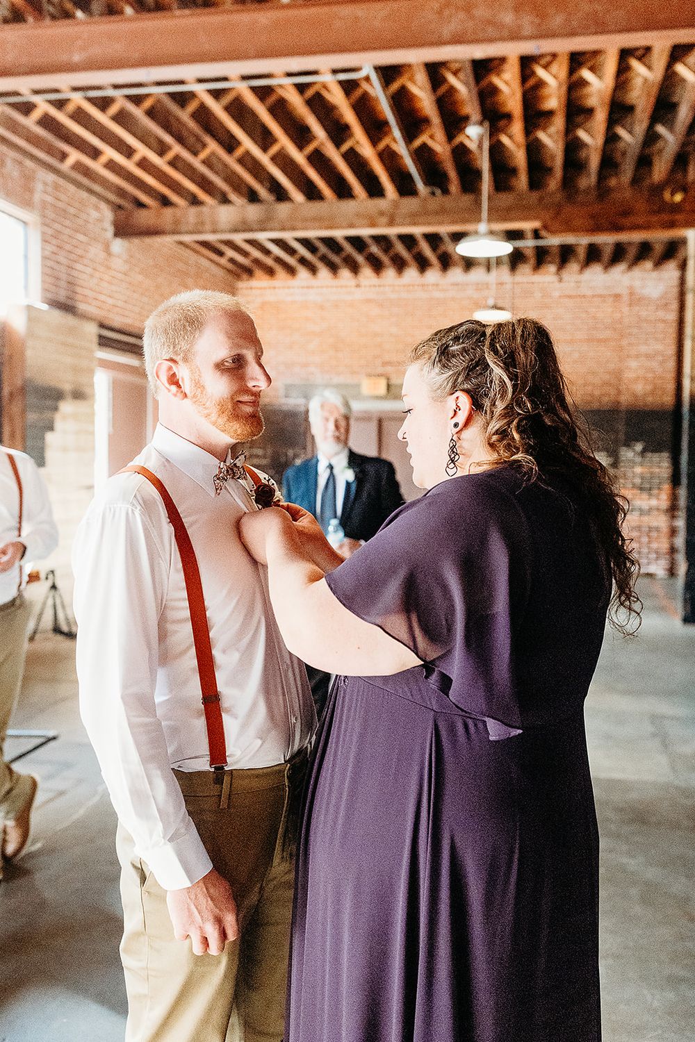 A person in purple helps adjust another's suspenders in a rustic brick venue with exposed wood beam ceiling.