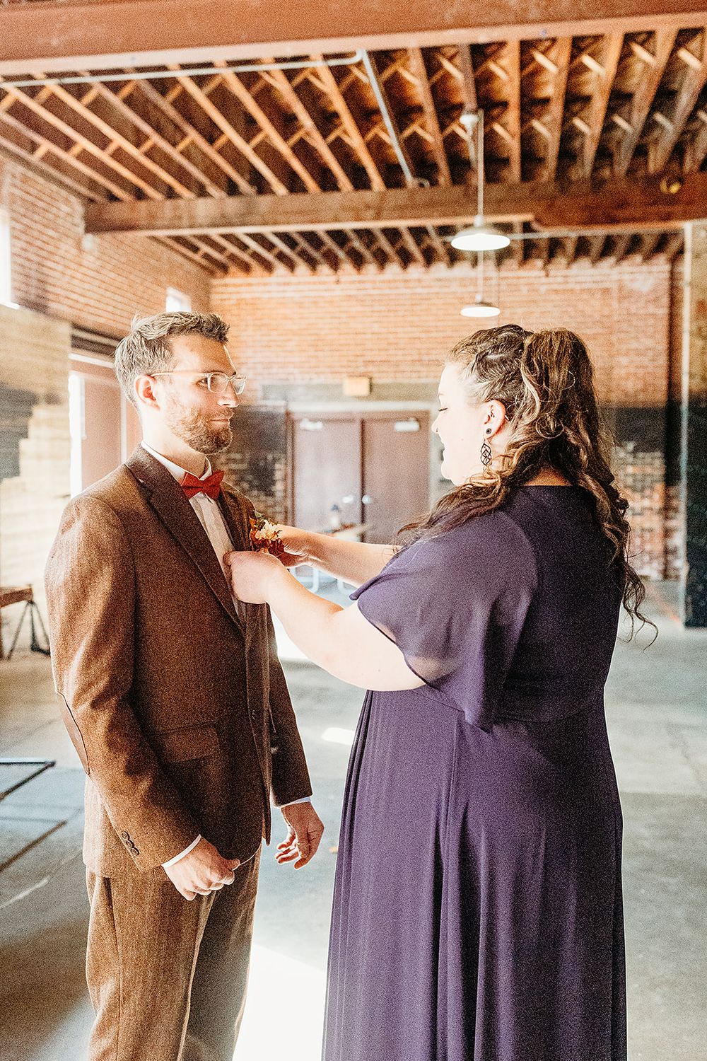 A person in a purple dress helps adjust a red bow tie on someone wearing a brown suit in an industrial brick interior space.