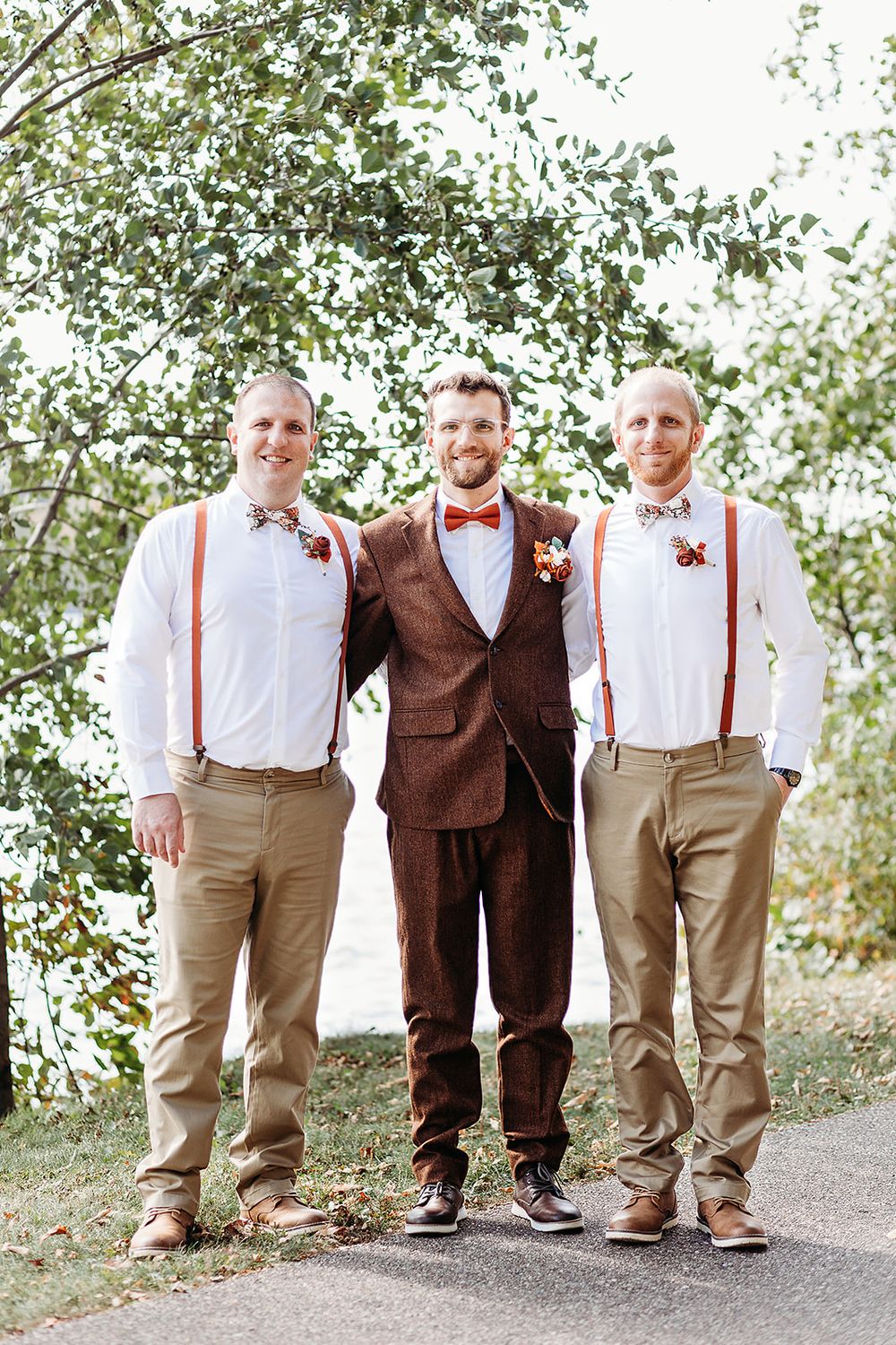 Three formally dressed attendants pose outdoors, two in bow ties and suspenders flanking one in a brown suit.