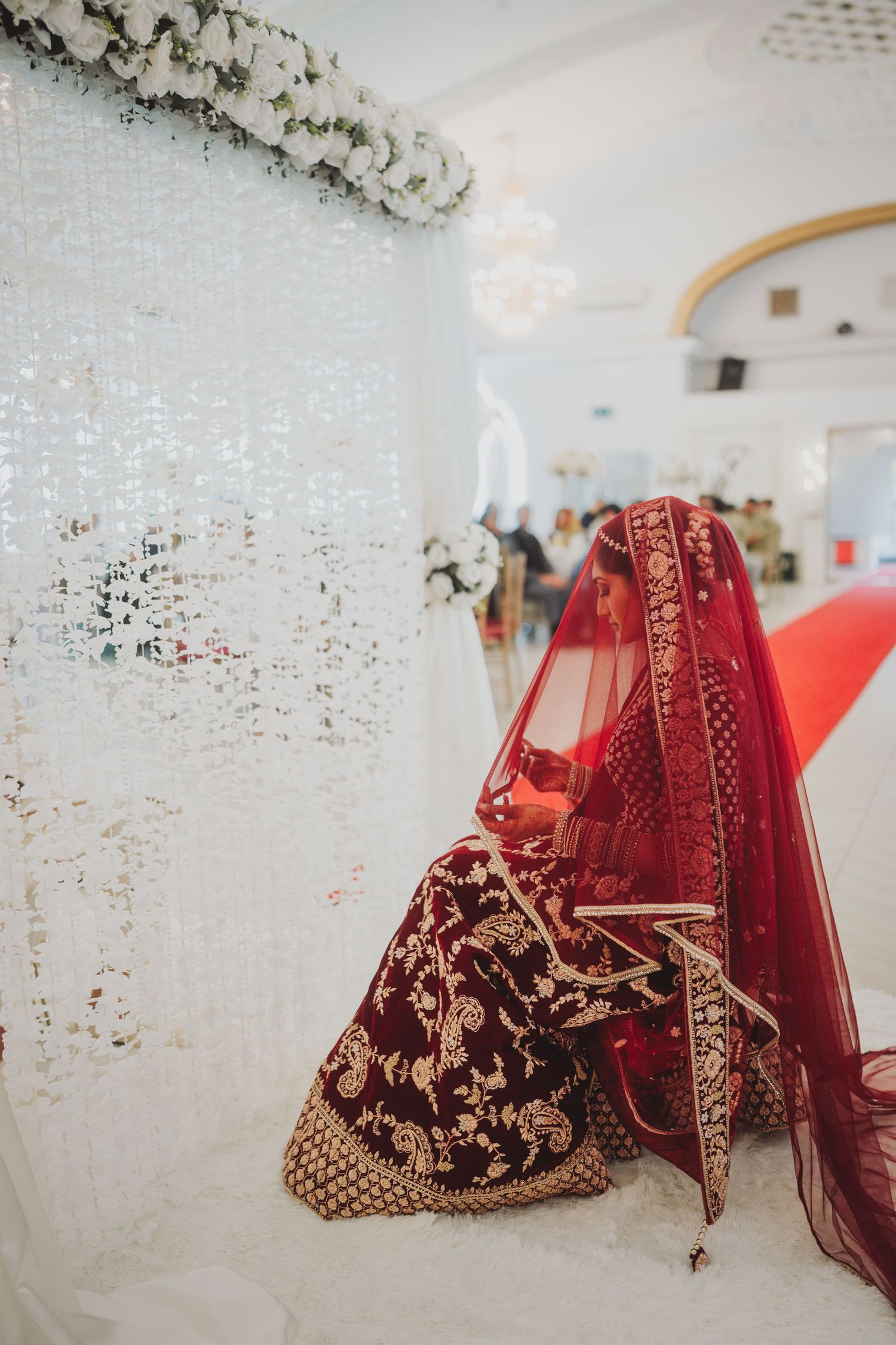 Elegant red and gold embroidered bridal lehenga displayed against white curtain backdrop.