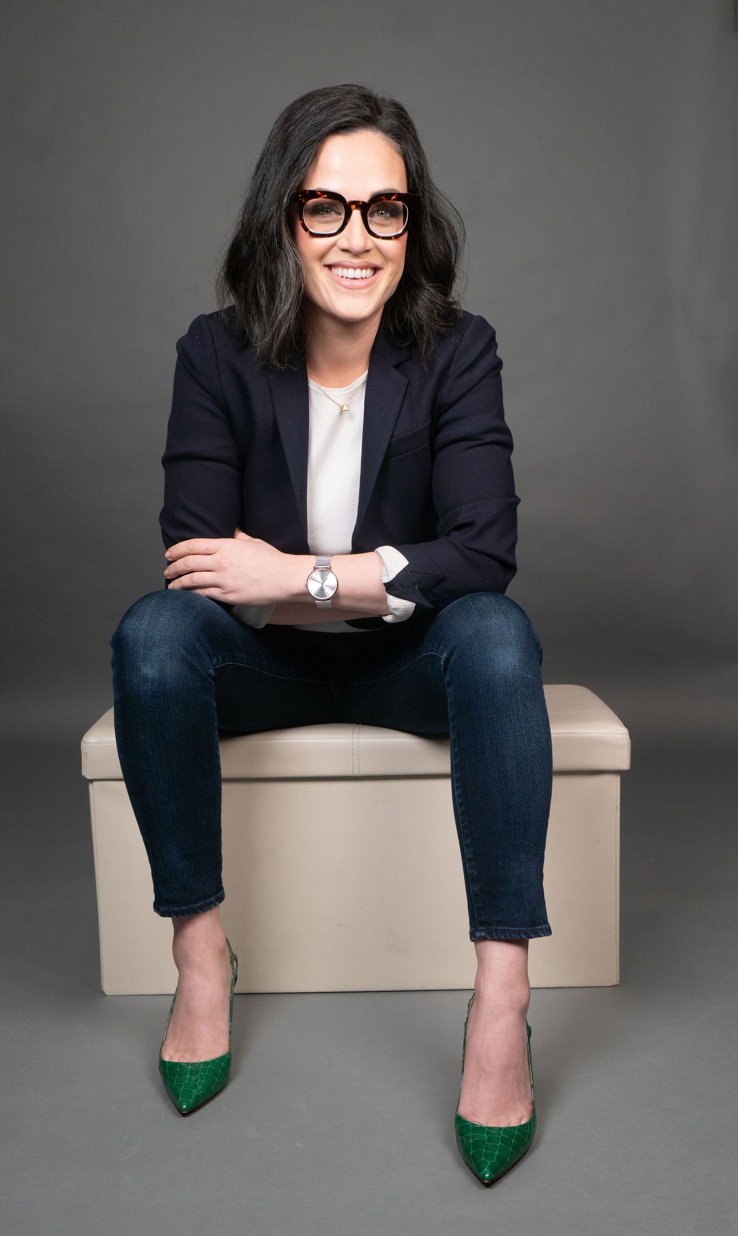 Woman in blazer and glasses sitting on beige ottoman wearing green shoes, smiling professionally at camera. Her arms are crossed.