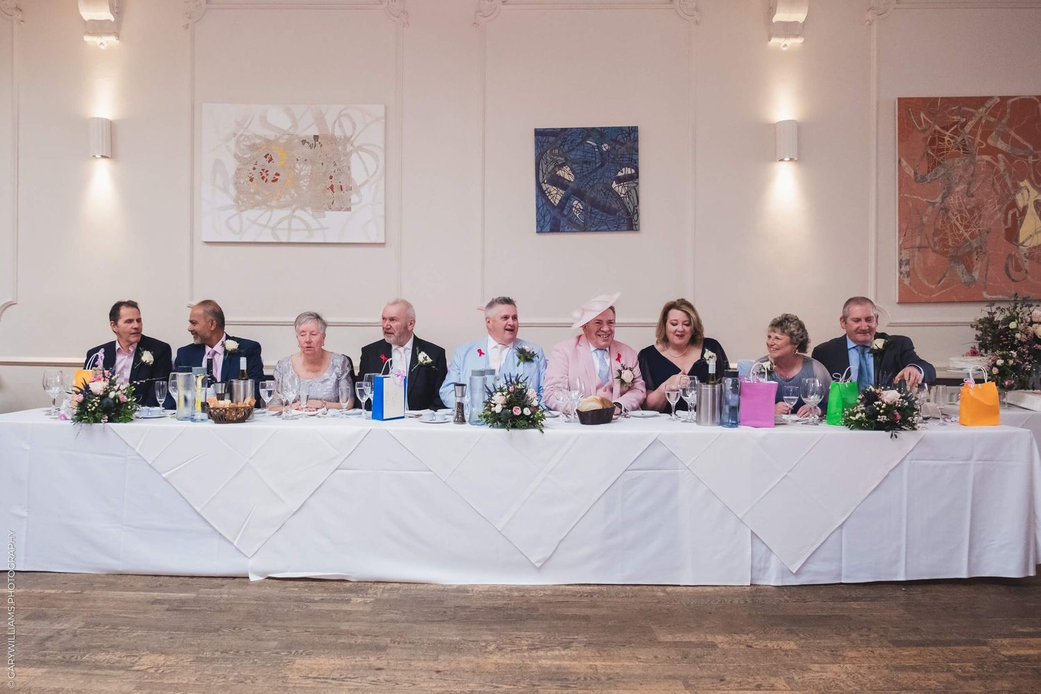 Wedding reception guests seated at a long white-clothed head table with floral centerpieces and drinks beneath wall artwork.