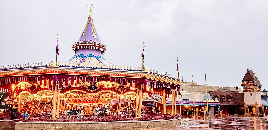 A colorful carousel with bright lights illuminates an amusement park pavilion under an overcast sky.