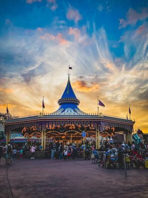Disney's Magic Kingdom carousel spins against vibrant sunset skies with flags waving and colorful lights glowing at dusk.