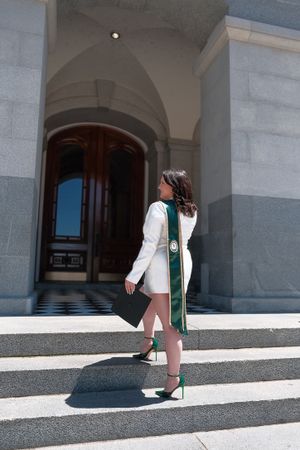Graduate in white dress and green sash standing on steps of elegant building entrance in graduation photo.
