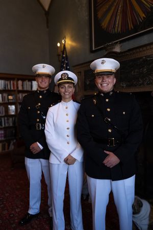 Three military service members in dress uniforms stand together indoors, wearing white caps and formal attire.