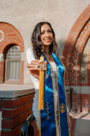 Graduate student poses at campus building wearing blue graduation stole and moving gold tassel in photo series.