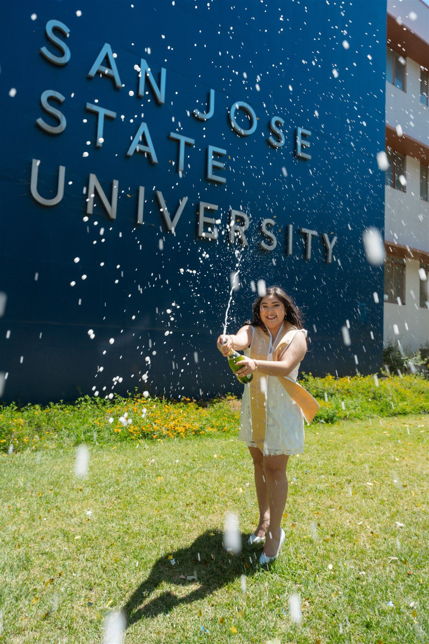 Person in white outfit stands on sunny grass with soap bubbles floating in front of San Jose State University building.