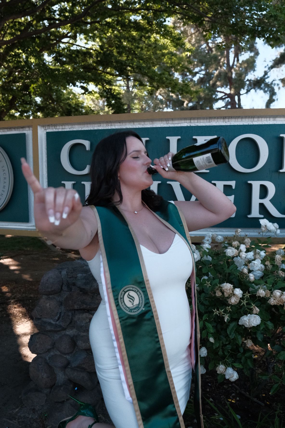 A graduate in white dress with green sash drinking from a bottle during outdoor celebration near flower bushes and signage.