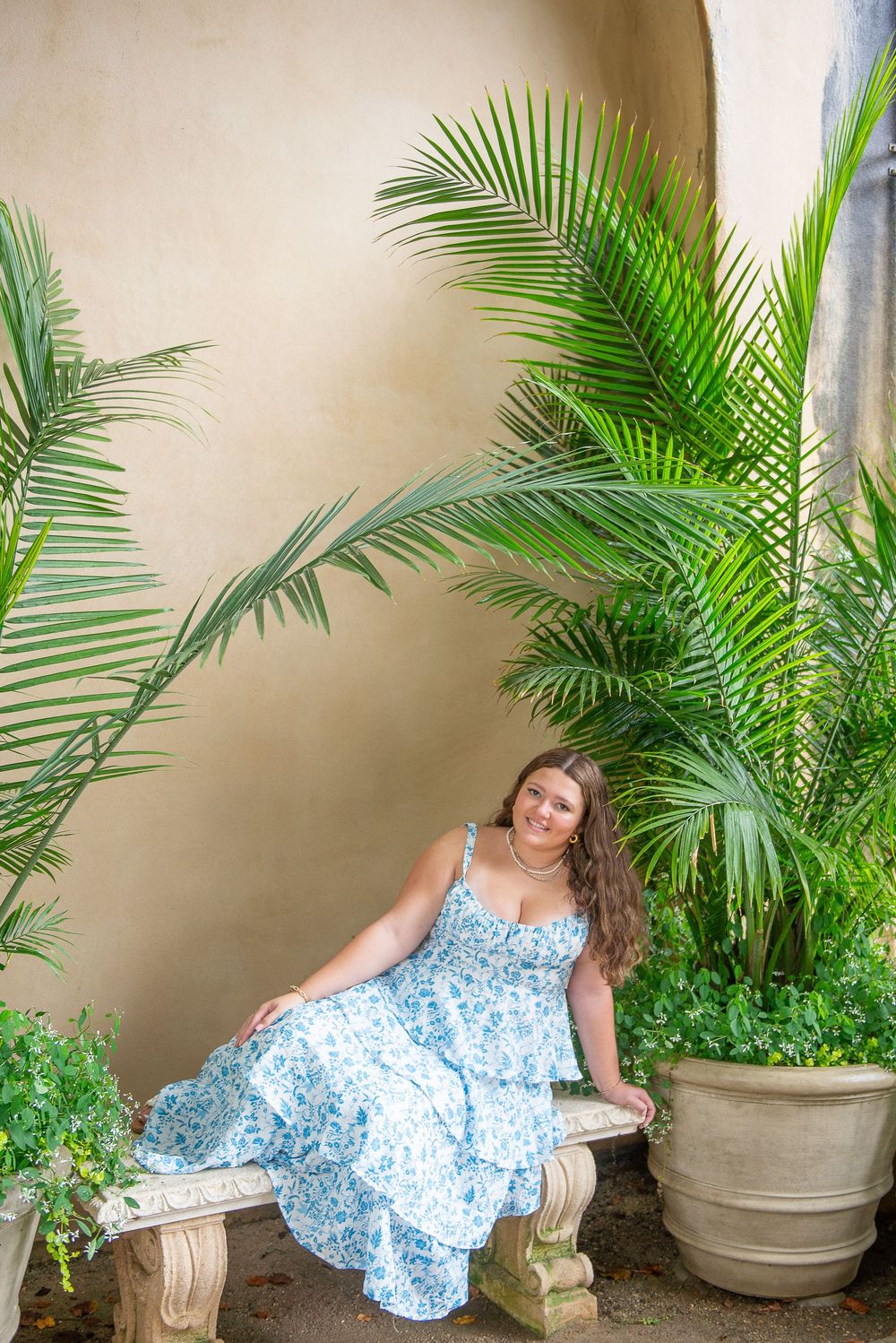 Senior portrait in a blue floral dress on a stone bench surrounded by tropical palm plants.