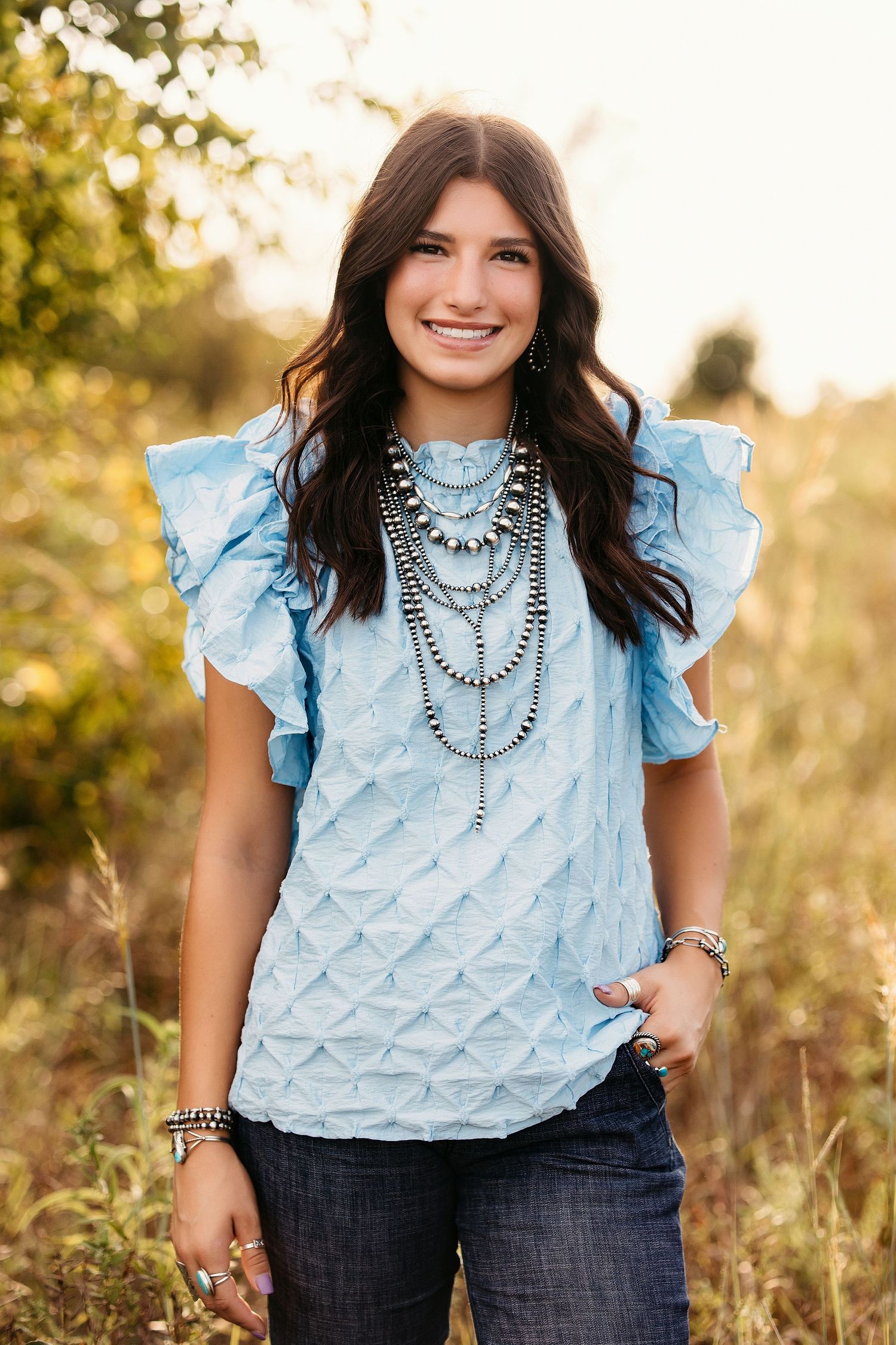 Woman wearing light blue ruffled top and statement necklace poses in outdoor setting with golden sunlight.