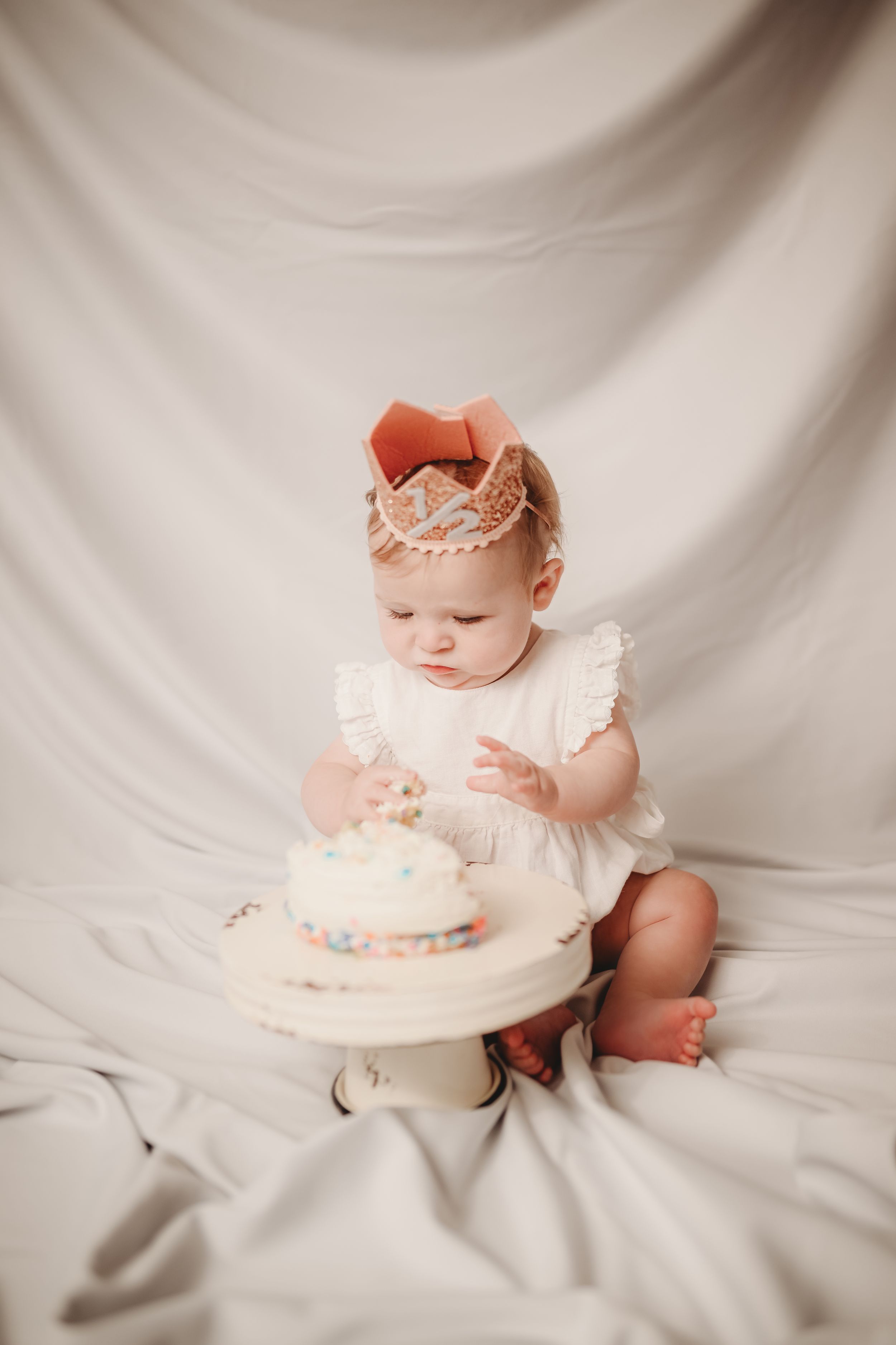 Baby in white dress having cake smash photo session on light backdrop.